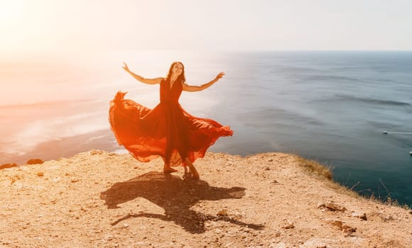 Side view a Young beautiful sensual woman in a red long dress posing on a rock high above the sea during sunrise. Girl on the nature on blue sky background. Fashion photo.