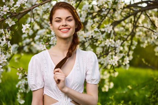 portrait of a joyful woman in a light dress against the background of a flowering tree, holding herself by her braid. High quality photo