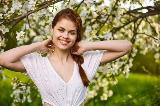 portrait of a joyful woman in a light dress against the background of a flowering tree, raising her hands to her head. High quality photo
