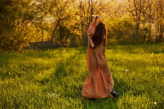 a slender woman with long hair stands in a field with her back to the camera, illuminated by the rays of the setting sun and joyfully raises her hands up. Horizontal photography on the theme of unity with nature. High quality photo