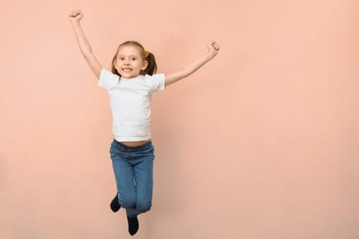Cute happy emotional caucasian girl 7 years old in a white t-shirt jumping on a pink background.