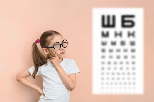 Thoughtful smart girl of elementary school age in round glasses against the background of a blurred sivtsev table for checking visual acuity.