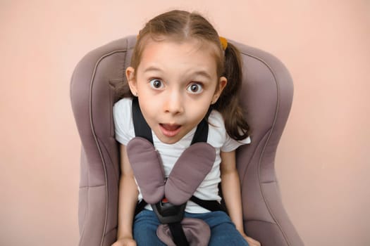 Funny surprised Caucasian girl of primary school age in a car seat, studio portrait.