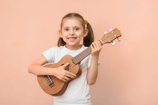 Happy funny caucasian smiling girl 6 - 8 years old in a white t-shirt with a ukulele on a pink background.