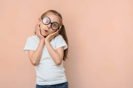 Cute surprised little caucasian girl in round glasses and hands near her face on a pink background with empty space.