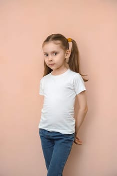 Caucasian girl 7 years old in a white T-shirt and jeans on a pink background, vertical studio portrait.