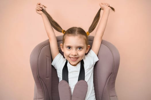 Funny happy caucasian girl with 2 ponytails of primary school age sits in a car seat, looks at the camera.