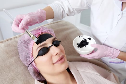 Cosmetologist applying black mask on the face of a beautiful woman for carbon peeling in beauty salon