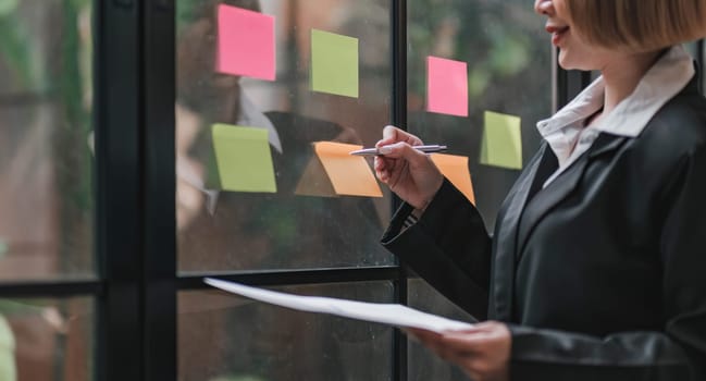 Young businesswoman creative team using post it notes in glass wall to writing strategy business plan to development grow to success...