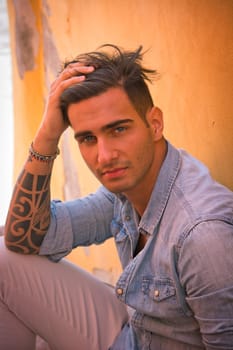 Portrait of handsome man with tattoo on arms sitting on cement stairs while leaning on wall, looking at camera