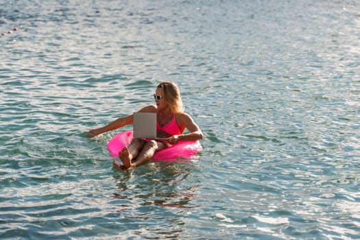 Woman works on laptop in sea. Freelancer, blond woman in sunglases floating on an inflatable big pink donut with a laptop in the sea at sunset. People summer vacation rest lifestyle concept