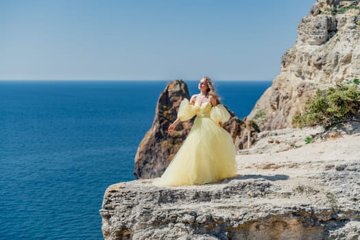 Woman in a yellow dress on the sea. Side view Young beautiful sensual woman in yellow long dress posing on a rock high above the sea at sunset. Girl in nature against the blue sky.