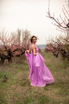 Woman peach blossom. Happy curly woman in pink dress walking in the garden of blossoming peach trees in spring.