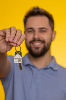 Happy young man real estate agent lifting hand showing the keys of new home house apartment, buying renting property, mortgage loan. Handsome guy isolated alone on yellow studio background. Vertical