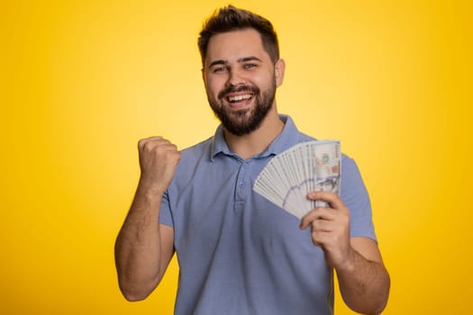 Rich pleased boss man waving money dollar cash banknotes bills like a fan, success business career, lottery winner, big income, wealth. Handsome caucasian guy isolated on yellow background, indoor