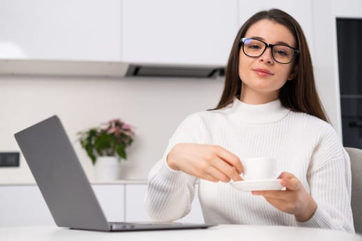 Portrait of a young beautiful woman holding a coffee cup and sitting in the kitchen near the laptop. Freelancer has a break