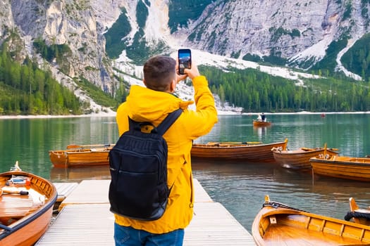 Tourist taking photo of wooden boats and Dolomites on the lake Braies. Man wearing yellow jacket and holding a black backpack visiting Dolomites Alps, Italy.