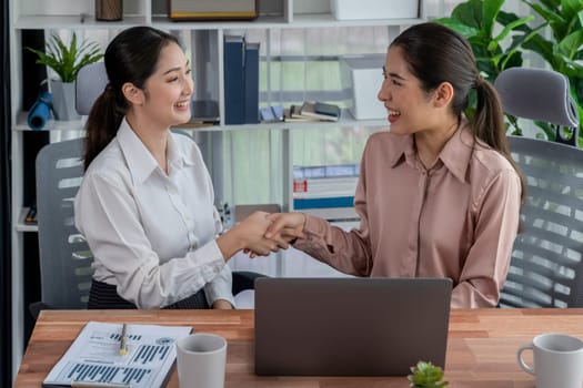 Two Asian businesswomen shaking hands in modern office after successfully working or analyzing using laptop as importance of teamwork and integrity in the workplace concept. Enthusiastic