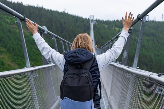 Female tourist with a backpack raised her hands and enjoys the walk on the longest suspension bridge Sky Bridge 721, Dolni Morava, Czech Republic