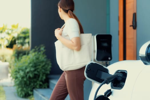 Focus image of electric vehicle recharging battery at home charging station with blurred woman walking in the background. Progressive concept of green energy technology applied in daily lifestyle.