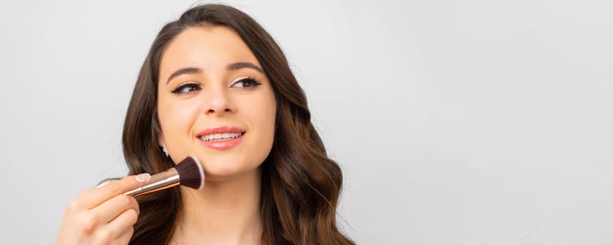 Young woman with perfect skin holding makeup brush near her face on the grey background with copy space