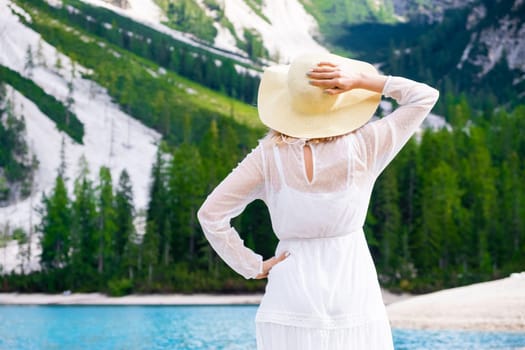 Young beautiful woman in a white dress holding a hat on the head against Lake Braies and mountains.