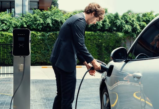 Progressive businessman with electric car recharging at public charging station at modern city residential area. Eco friendly rechargeable car powered by alternative clean energy.