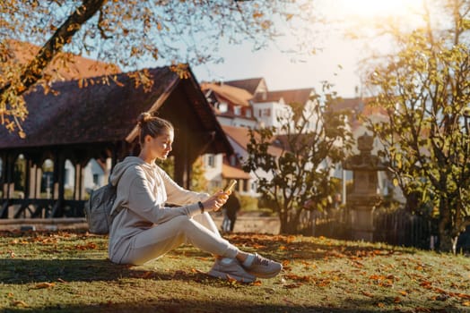 Young fashionable teenage girl with smartphone in park in autumn sitting at smiling. Trendy young woman in fall in park texting. Retouched, vibrant colors. Beautiful blonde teenage girl wearing casual modern autumn outfit sitting in park in autumn. Retouched, vibrant colors, brownish tones.