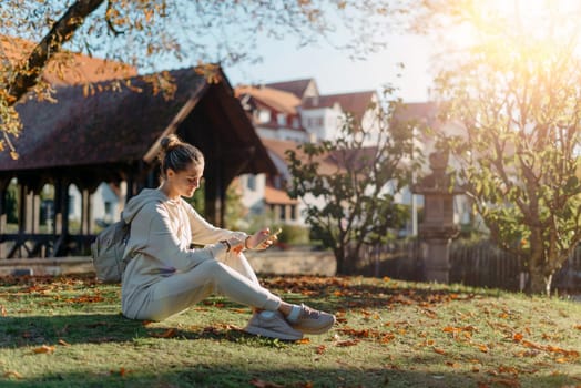 Young fashionable teenage girl with smartphone in park in autumn sitting at smiling. Trendy young woman in fall in park texting. Retouched, vibrant colors. Beautiful blonde teenage girl wearing casual modern autumn outfit sitting in park in autumn. Retouched, vibrant colors, brownish tones.