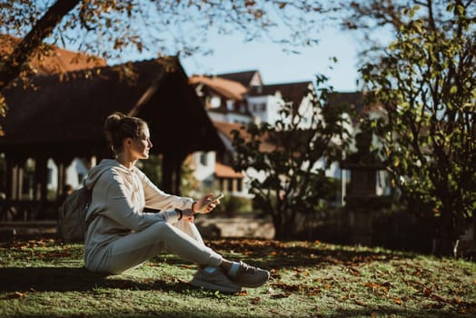 Young fashionable teenage girl with smartphone in park in autumn sitting at smiling. Trendy young woman in fall in park texting. Retouched, vibrant colors. Beautiful blonde teenage girl wearing casual modern autumn outfit sitting in park in autumn. Retouched, vibrant colors, brownish tones.