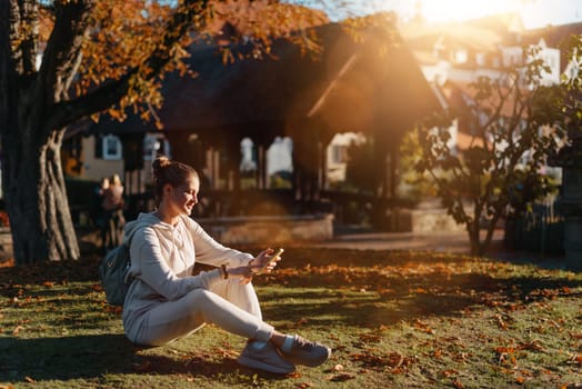 Young fashionable teenage girl with smartphone in park in autumn sitting at smiling. Trendy young woman in fall in park texting. Retouched, vibrant colors. Beautiful blonde teenage girl wearing casual modern autumn outfit sitting in park in autumn. Retouched, vibrant colors, brownish tones.