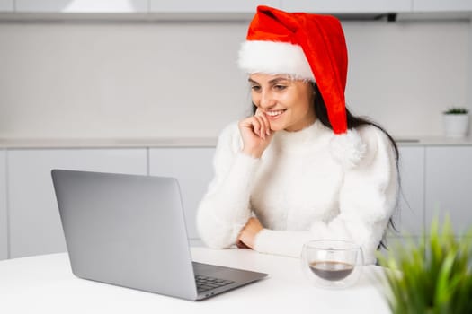 A woman in red Santa Claus watches a film or video and smiles using her laptop during the Christmas holidays.