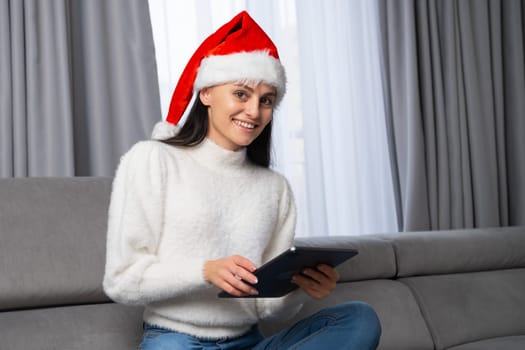 Christmas portrait of a beautiful girl wearing a Santa Claus hat and sitting on the sofa with a tablet in her hand in a modern home interior. Ordering of gifts via the internet on Christmas or New Year.