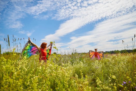 Happy female family with red haired mother and daughter with bright butterfly wings having fun on green and yellow meadow full of grass and flowers in sunny summer day. Concept family love