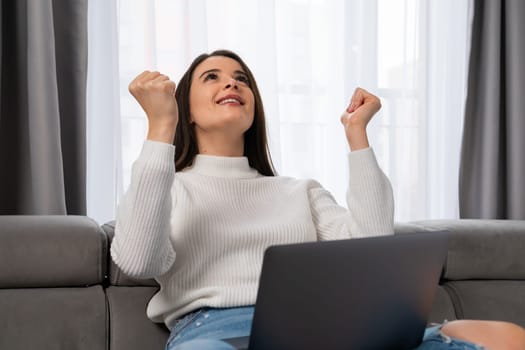 An excited young woman rejoices in internet victory holding a laptop on her lap in a cozy living room.