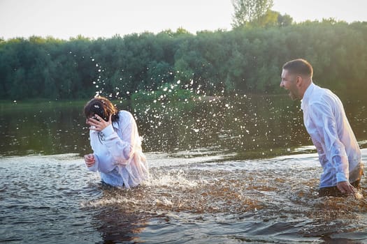 Beautiful adult couple has fun in nature in the water in a river or lake in the summer evening at sunset. A guy and a girl swim and relax outdoors in clothes in white shirts and jeans