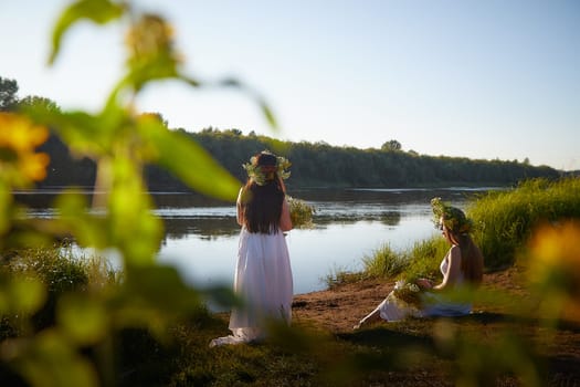 Adult mature and young brunette women in white sundresses and wreaths of flowers in summer by water of river or lake at sunset. Girl mother and daughter Celebrating pagan holiday of Ivan Kupala