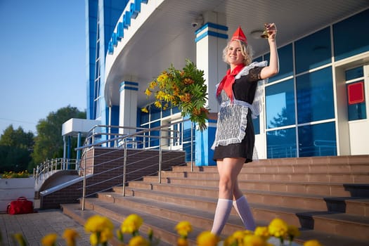 A Girl in black uniform, white apron, red tie on steps of school with bouquet of flowers and bell. Nostalgia photo shoot of teenager of female pioneer from USSR costume for September 1 or graduation