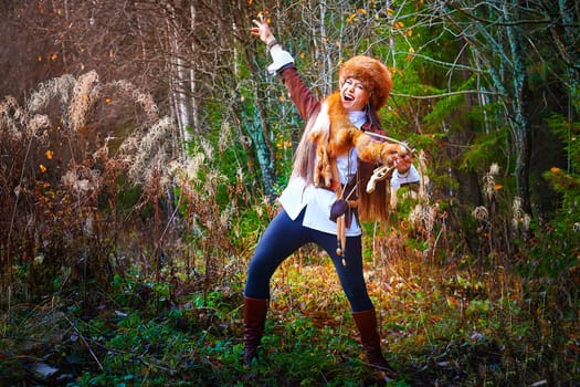 Girl in a leather jacket, a big red fox fur hat and with the skin of a fox killed on the hunt in the forest in autumn. A female model poses as fabulous royal huntress on nature hunt at photo shoot