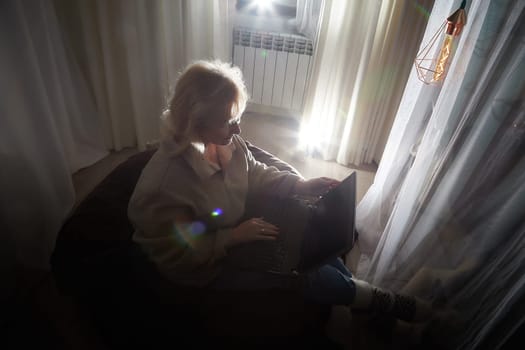 Silhouette and shadow of Adult mature woman of 40-60 years with laptop, notebook, computer in warm sweater in dark calm cozy evening atmosphere room. Interior with curtains and small linght