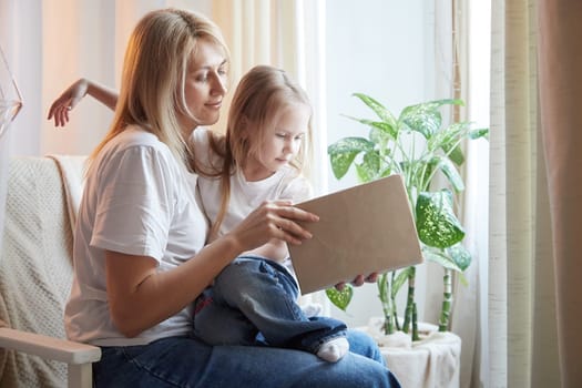 Happy loving family with mother and daughter reading book in living room. Woman mom and small child girl having convercation and studing inside of home