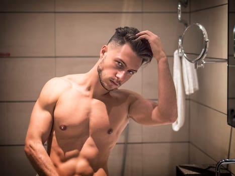 Shirtless muscular handsome young man looking at camera in bathroom in the morning, wearing only white towel around waist