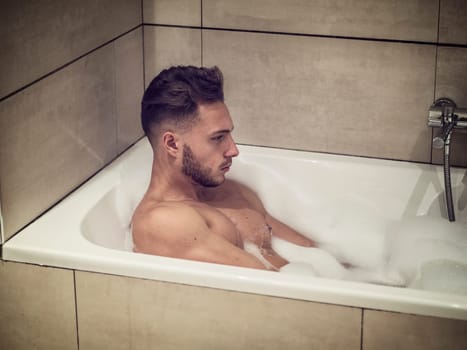 Handsome young man in bathtub at home having bath, leaning on tub edge,