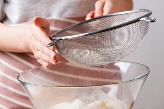 Close up woman sifts wheat flour through a sieve into mixing bowl for dough preparation for cake and bakery on the black background. Knead the dough for homemade pastry.