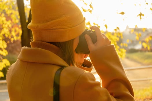 A woman in an orange hat and brown coat photographs the autumn park, back view.