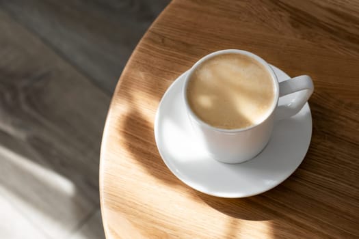 A white porcelain cup with coffee on a saucer stands on a small round table made of natural wood. Morning, coffee, hot drink. Hard light.