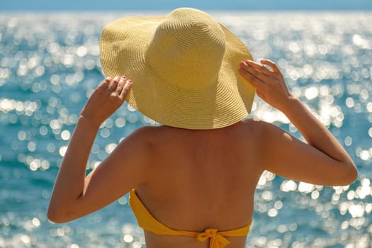 Woman in yellow bikini and straw hat looks at blue sea on warm day. Lady holds headdress with hands against water sparkling in sun close back view