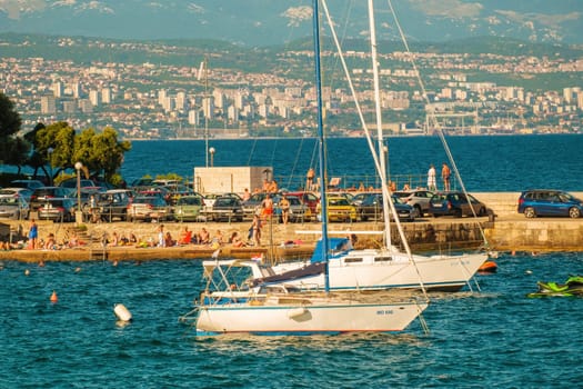 White boats drift on deep turquoise water near sea pier with people and cars on sunny day. Coastal city resort against high mountains