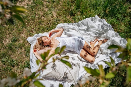 woman sleeps on a white bed in the fresh spring grass in the garden. Dressed in a blue nightgown