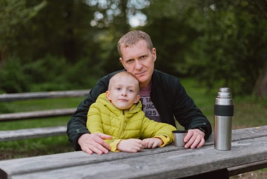 Happy family: father and child boy son playing and laughing in autumn park, sitting on wooden bench. Father and little kid having fun outdoors, playing together. Father and son sitting on a bench and talking. dad son park bench table autumn thermos.